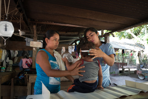 Two women measure a child's head in a rustic outdoor care setting
