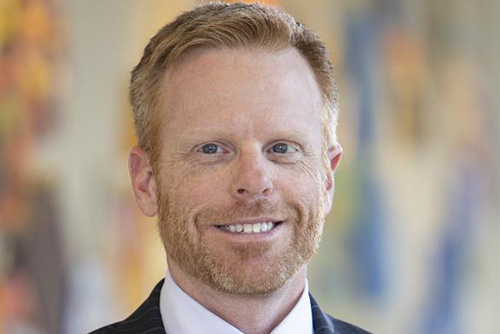 A portrait of Joel Stoddard wearing a dark pinstripe business suit and floral tie