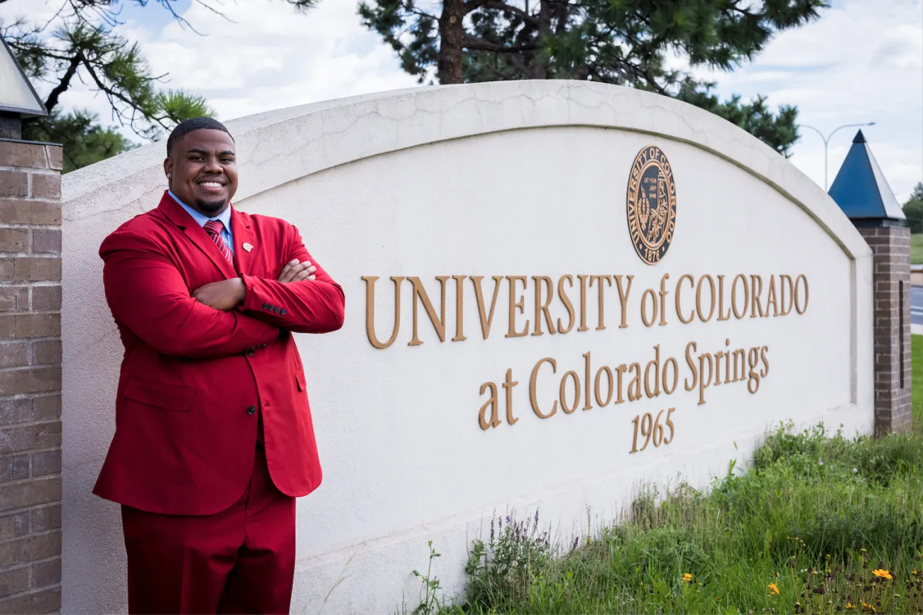 Michael "Axel" Brown wears a red suit and poses next to the University of Colorado Colorado Springs sign